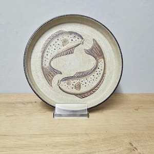 May include: A round, beige ceramic plate with a dark brown rim. Two stylized brown fish are depicted in the center, swimming in a circle. The fish have dotted patterns and detailed fins. The plate is displayed on a wooden surface.