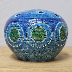 May include: A ceramic vase with a textured, spherical shape. The vase is primarily blue with darker blue and green accents. It features three large, circular designs with green centers and several small holes on top.