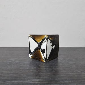 Può includere: Un piccolo vaso quadrato in ceramica con un design astratto in marrone, nero e bianco. Il vaso è appoggiato su una superficie di legno scuro.