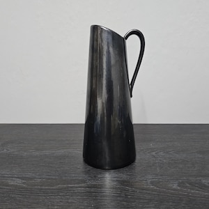 Könnte beinhalten: Eine schwarze Keramikkanne mit einem geschwungenen Henkel. Die Kanne hat eine glatte, glänzende Oberfläche.
