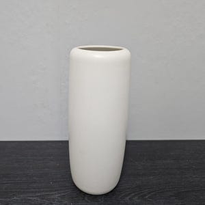 Könnte beinhalten: Eine weiße Keramikvase mit abgerundeter, zylindrischer Form. Die Vase ist glatt und hat eine matte Oberfläche.