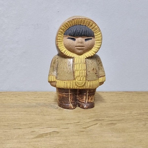 Könnte beinhalten: Eine Keramikfigur einer Person in einem Kapuzenparka. Die Figur hat schwarzes Haar und ist in Braun- und Beigetönen gekleidet. Der Parka hat eine gelbe Kapuze und einen gelben Besatz. Die Figur steht auf einer Holzoberfläche.