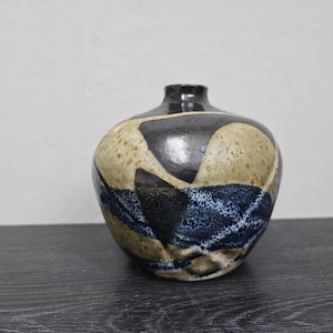 Può includere: Un piccolo vaso rotondo in ceramica con una smaltatura marrone e nera macchiata. Il vaso ha un motivo geometrico blu e bianco.