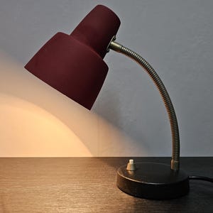 Könnte beinhalten: Eine Vintage-Tischlampe mit einem maronenfarbenen Schirm und einem flexiblen Metallarm. Die Lampe hat einen schwarzen Sockel und einen weißen Schalter.
