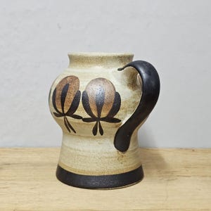 May include: A ceramic mug with a beige body, black handle, and a black band at the base. The mug features two floral designs in brown and black. The mug is sitting on a wooden surface.