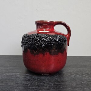 Könnte beinhalten: Eine rote Keramikvase mit einem schwarzen, strukturierten Rand und einem Henkel. Die Vase hat eine glänzende Oberfläche.