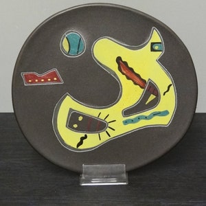 Peut inclure: Une assiette en céramique avec un fond brun foncé et un motif abstrait jaune. Le motif présente une grande forme jaune avec des lignes noires et rouges, un cercle bleu et un rectangle rouge avec des points jaunes.