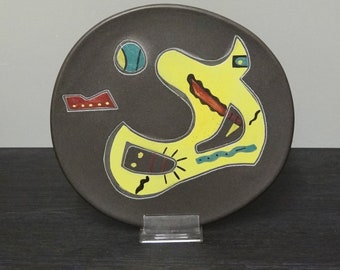 May include: A ceramic plate with a dark brown background and a yellow abstract design. The design features a large yellow shape with black and red lines, a blue circle, and a red rectangle with yellow dots.