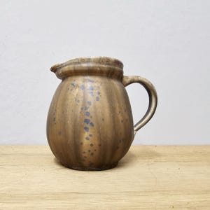 May include: A brown ceramic pitcher with a handle and spout. The pitcher has a bulbous shape with vertical ridges and blue speckles. It sits on a wooden surface against a neutral background. The pitcher is likely used for serving beverages.