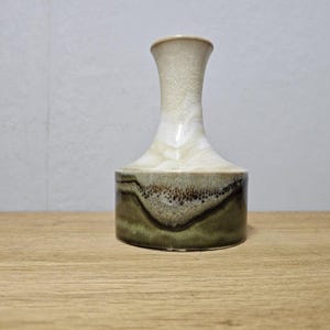 Könnte beinhalten: Eine Keramikvase mit zylindrischem Sockel und ausgestelltem Hals. Die Vase weist einen Farbverlauf auf, der von hellem Beige oben zu dunklem Olivgrün am Boden übergeht. Die Vase ist etwa 18 cm hoch.