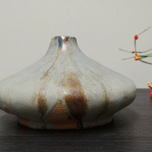 Könnte beinhalten: Eine kleine, graue Keramikvase mit einem breiten, abgerundeten Boden und einem schmalen Hals. Die Vase hat eine gefleckte Glasur mit braunen und blauen Akzenten.