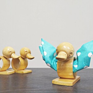 May include: Wooden duck-shaped napkin rings. Two rings are side-by-side, and a third duck holds a blue napkin with white polka dots. The ducks are light brown with carved details, and sit on small wooden bases.