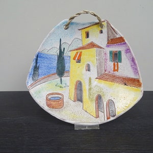 Puede incluir: Un colgante de pared de cerámica con una escena pintada de una casa amarilla con techo rojo, un pozo y un mar azul en el fondo. La casa tiene una ventana verde y un toldo rojo. El colgante de pared tiene una manija de cuerda.