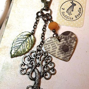 May include: A decorative keychain with a bronze-colored tree charm, a green leaf, and a heart-shaped charm with text and a sunflower design. The keychain also features a small orange bead and a tag with a rabbit logo.
