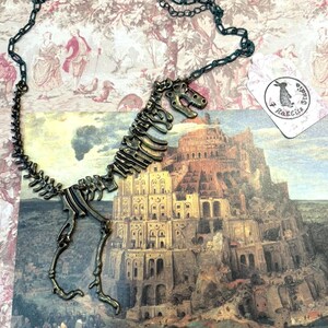May include: A bronze-colored dinosaur skeleton pendant necklace. The pendant is attached to a dark green chain. The necklace is displayed on a vintage-style background with a painting of a large building and a tag with a rabbit logo.