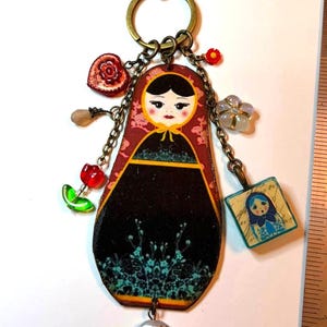 May include: A keychain featuring a flat, wooden matryoshka doll pendant with a black and red floral design, adorned with multiple charms including a red heart, a green tulip, a blue square doll charm, and a round floral bead, with a metal ruler visible.