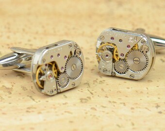 May include: A pair of silver cufflinks with exposed clockwork mechanisms. The cufflinks have a square shape and feature a variety of gears and other clock parts.