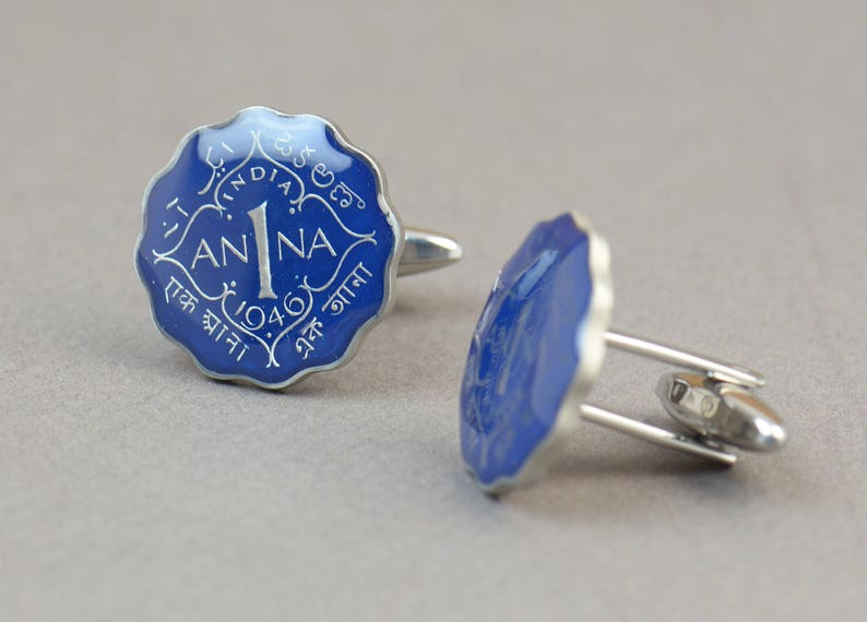May include: A pair of silver cufflinks with a blue enamel face. Each cufflink features a scalloped edge and the text "1 ANNA 1946" in white. The cufflinks have a classic, elegant design.