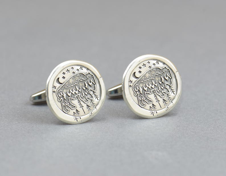 May include: A pair of silver cufflinks with a detailed design of a bison standing in a forest with stars and a crescent moon in the background.