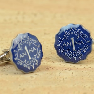 May include: A pair of cufflinks featuring a blue and silver design. Each cufflink has a scalloped edge and displays the word "ANNA" with the number "1" and the year "1946" in white lettering. The cufflinks are made of metal.