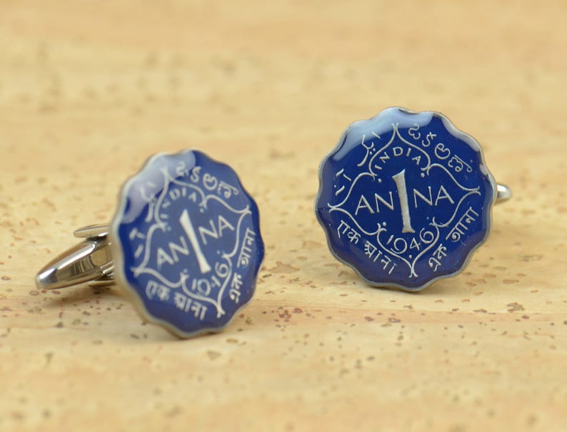 May include: A pair of cufflinks featuring a blue and silver design. Each cufflink has a scalloped edge and displays the word "ANNA" with the number "1" and the year "1946" in white lettering. The cufflinks are made of metal.