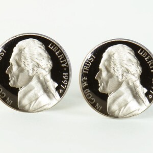 May include: Two silver-coloured cufflinks featuring a profile of a man's face with the words "In God We Trust" and "Liberty 1997" on each cufflink.