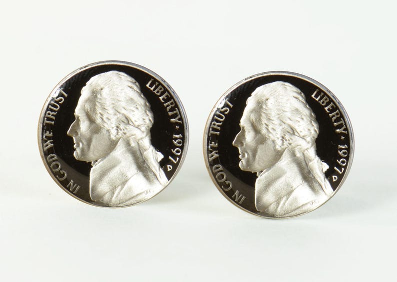 May include: Two silver-coloured cufflinks featuring a profile of a man's face with the words "In God We Trust" and "Liberty 1997" on each cufflink.