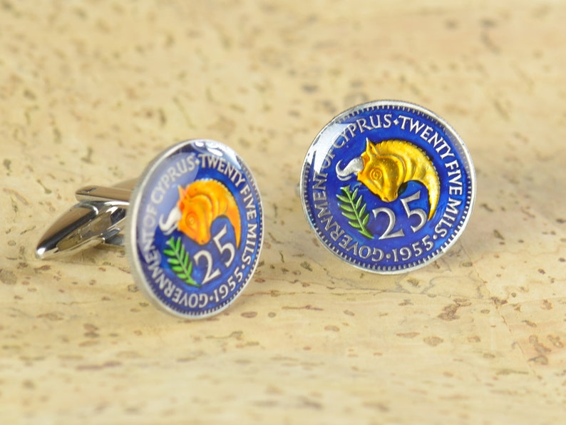 Peut inclure: Une paire de boutons de manchette repr&eacute;sentant une pi&egrave;ce de monnaie chypriote vintage de 1955 avec un fond bleu, un taureau dor&eacute; et le texte "GOVERNMENT OF CYPRUS TWENTY FIVE MILLS 1955-5 25".