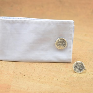 May include: A pair of silver cufflinks with a detailed design of a tree with a crescent moon and stars. The cufflinks are on a white shirt cuff.
