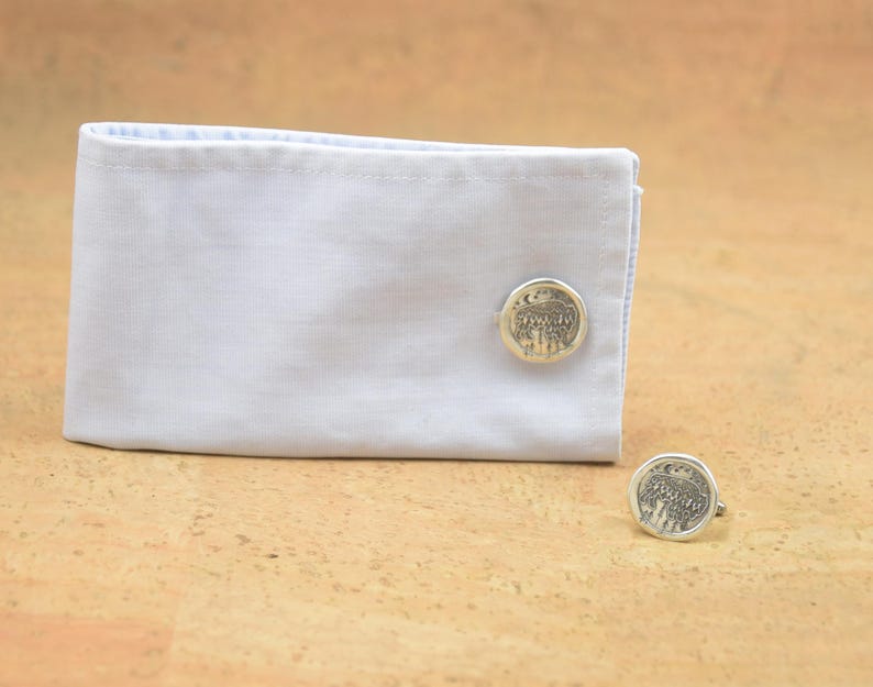 May include: A pair of silver cufflinks with a detailed design of a tree with a crescent moon and stars. The cufflinks are on a white shirt cuff.