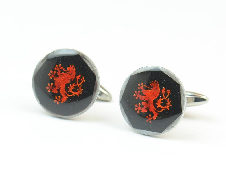 May include: A pair of silver cufflinks with a black background and a red lion crest design.