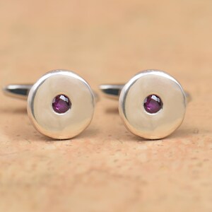 May include: A pair of silver cufflinks with a round, flat design. Each cufflink features a small, round, purple gemstone in the center.