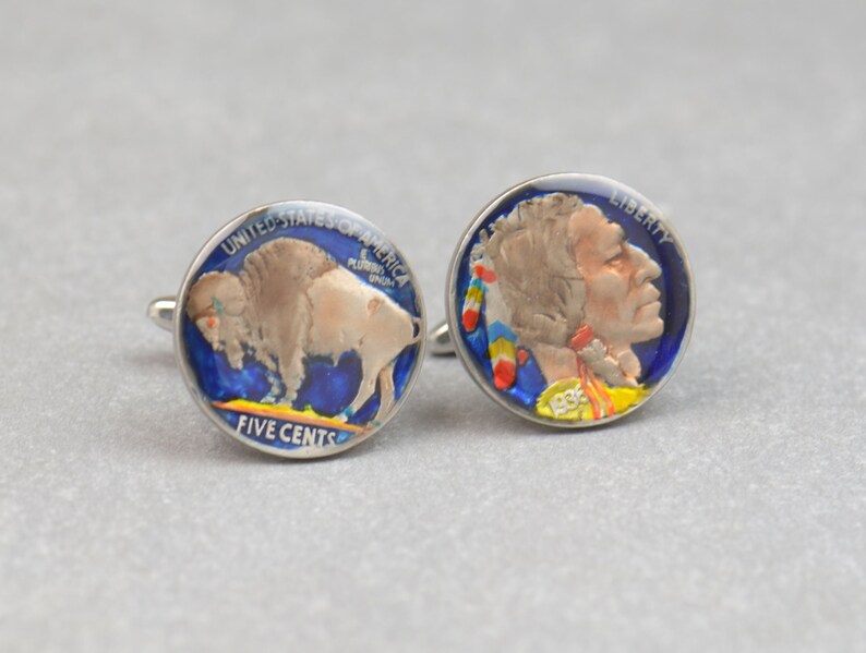 May include: A pair of cufflinks made from repurposed five-cent coins. One cufflink features a painted image of a buffalo, the other features a painted image of an American Indian. Both cufflinks have a blue background with yellow and red accents.