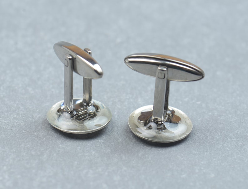 May include: A pair of silver cufflinks with a round, white, and blue design. The cufflinks have a silver oval-shaped bar with a small square in the center.