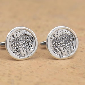 May include: A pair of silver cufflinks with a detailed design of a bison standing in a forest with a crescent moon and stars in the background.