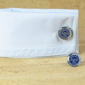 May include: A pair of silver cufflinks with a blue enamel design. The cufflinks feature the text "BRASIL 300 DEIS 1938".