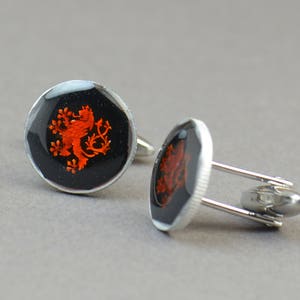 May include: A pair of silver cufflinks with a black background and a red lion crest design.