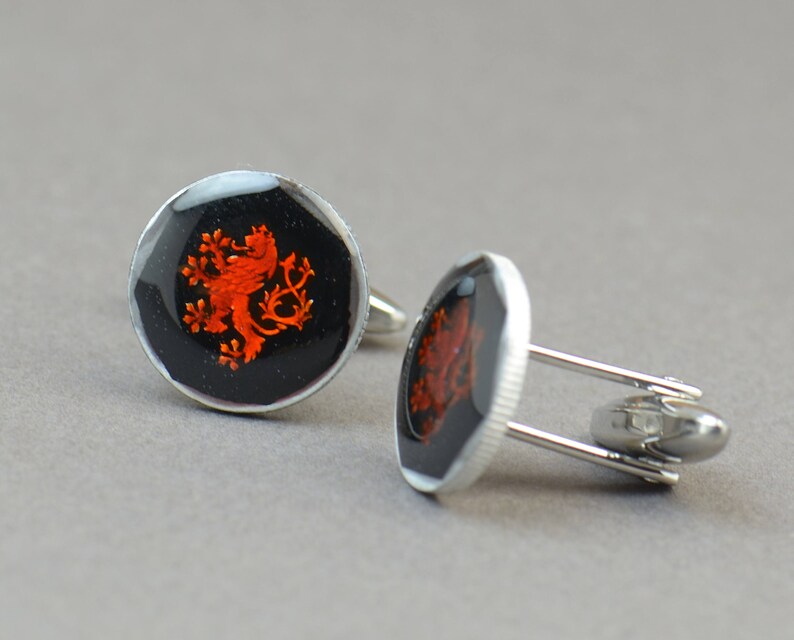 May include: A pair of silver cufflinks with a black background and a red lion crest design.