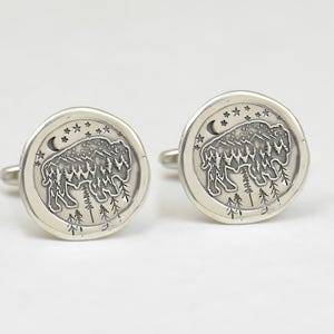 May include: A pair of silver cufflinks with a detailed design of a buffalo standing in a forest with a crescent moon and stars in the background.