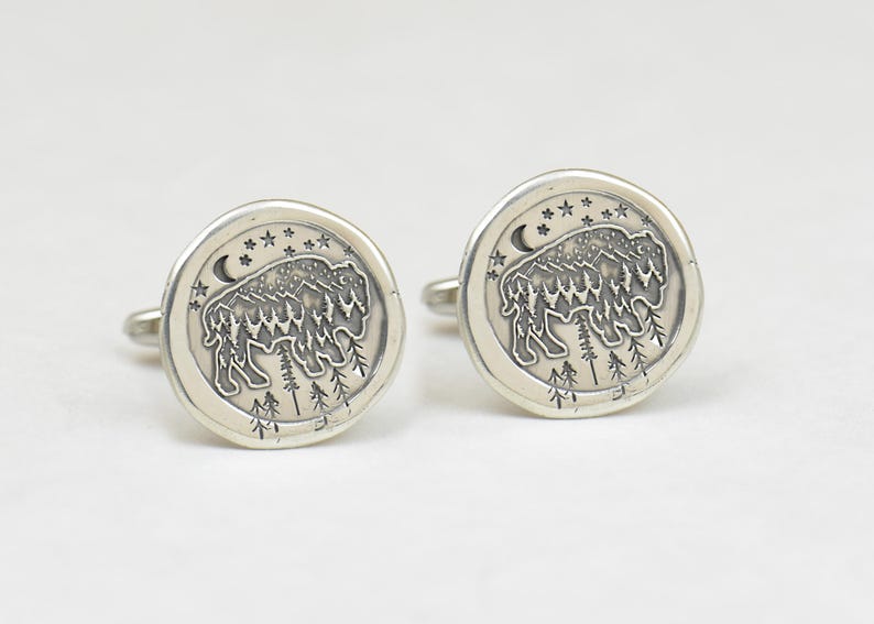 May include: A pair of silver cufflinks with a detailed design of a buffalo standing in a forest with a crescent moon and stars in the background.