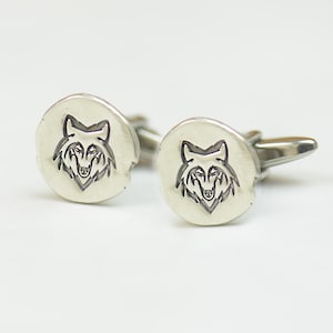 Op de afbeelding: Een paar zilveren manchetknopen met een wolfskopontwerp. De wolfskop is gegraveerd op een rond, getextureerd oppervlak.