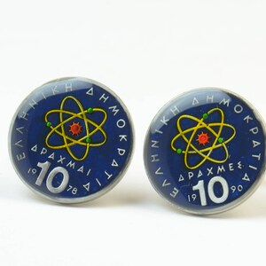May include: Two silver-colored cufflinks with a blue background and a yellow atom symbol. The text "ΕΛΛΗΝΙΚΗ ΔΗΜΟΚΡΑΤΙΑ" is written around the edge of the cufflinks. The text "10 ΔΡΑΧΜΕΣ" is written below the atom symbol. The year "1978" is written below the text "10 ΔΡΑΧΜΕΣ" on one cufflink and the year "1990" is written below the text "10 ΔΡΑΧΜΕΣ" on the other cufflink.