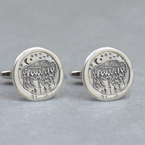 May include: A pair of silver cufflinks with a design of a bison standing in a forest with a crescent moon and stars in the background.
