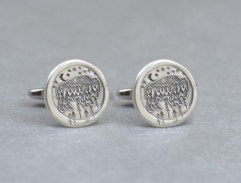 May include: A pair of silver cufflinks with a design of a bison standing in a forest with a crescent moon and stars in the background.