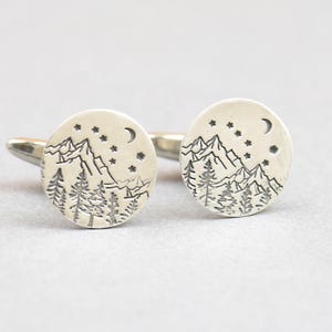 Può includere: Un paio di gemelli d'argento con un design di una catena montuosa, alberi, una luna crescente e stelle.