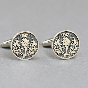 May include: A pair of silver cufflinks, each with a circular face. The design features a thistle emblem, a stylized floral motif, in a lighter tone set against a textured, darker background. The cufflinks are silver-toned.