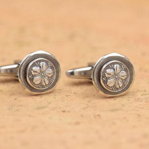 Può includere: Un paio di gemelli d'argento. Ogni gemello presenta un design circolare con un motivo floreale in rilievo al centro. I gemelli hanno un aspetto classico ed elegante, adatti per occasioni formali.