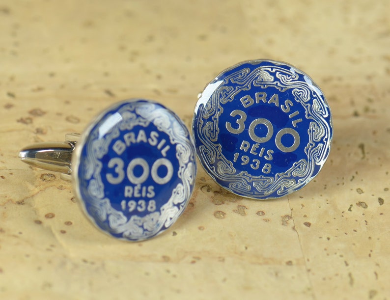 May include: A pair of cufflinks featuring a blue enamel design with silver accents. The cufflinks are round and feature the text "BRASIL 300 REIS 1938".