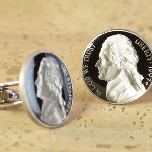 May include: A pair of silver cufflinks featuring a black and white image of a nickel coin with the profile of Thomas Jefferson on one side and the words "IN GOD WE TRUST" and "LIBERTY 1997" on the other side.