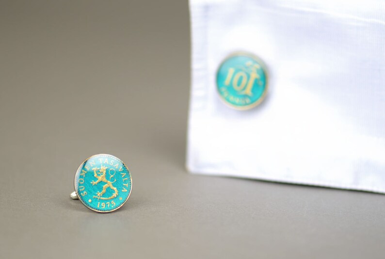 Pode incluir: Um par de bot&otilde;es de punho com uma moeda azul-esverdeada e dourada com um desenho de le&atilde;o. A moeda diz "SUOMEN TASAVALTA 1975".